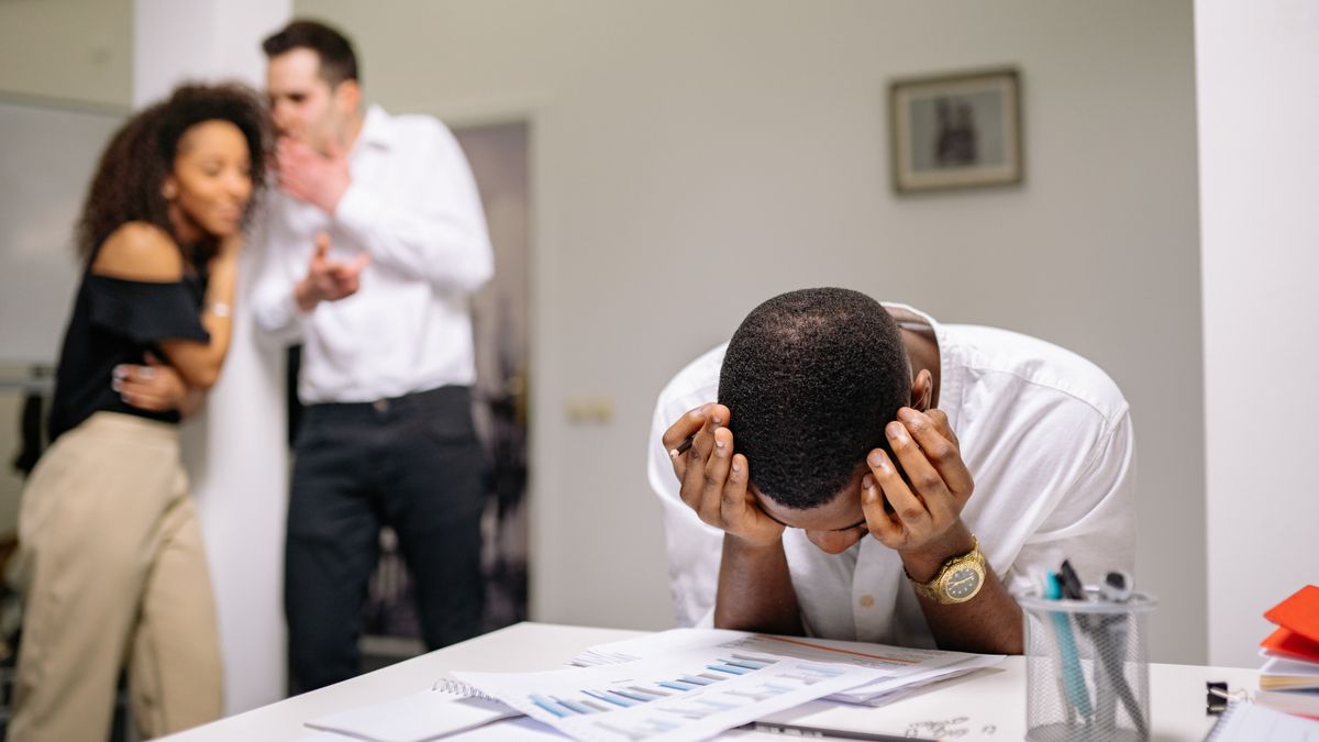 Trabalhador com as mãos na cabeça enquanto colegas conversam ao fundo, ilustrando assédio moral no trabalho